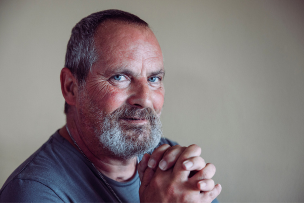 A portrait of a middle-aged man with light gray hair and a short gray beard. He has blue eyes and is looking to the right of the camera. His hands are clasped together in prayer.