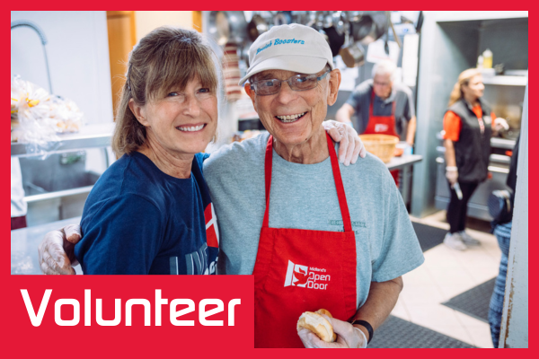 Two smiling volunteers, a woman and a man, in a kitchen setting. The man wears a red apron with "Open Door" logo. Text overlay reads "Volunteer".