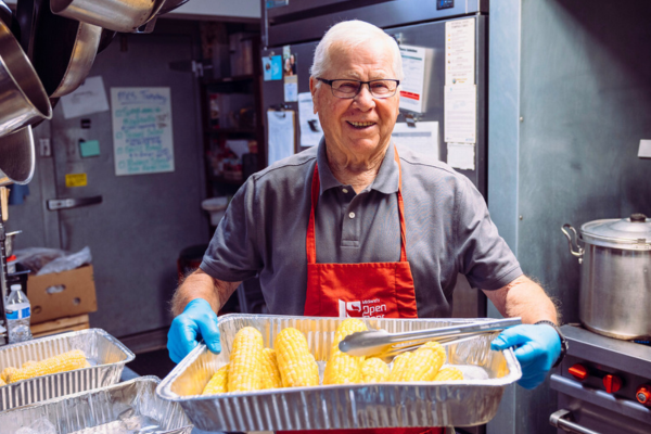 Volunteer – Food Service A smiling man holds a steaming tray of corn on the cob at Midland's Open Door's community soup kitchen.