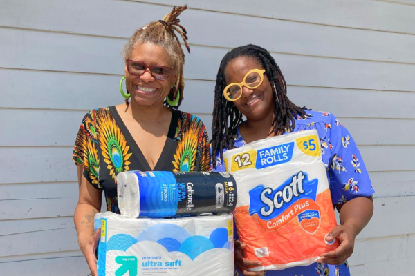 Donate – Personal Care Products Two women representing Breaking Bread Village stand with arms full of donations including paper towels and toilet paper for the Open Door.