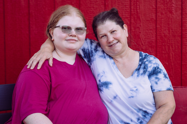 A mother and daughter sit on a bench in front of Open Door's clothing shed; the mother has her arm around her daughter with a wide smile.